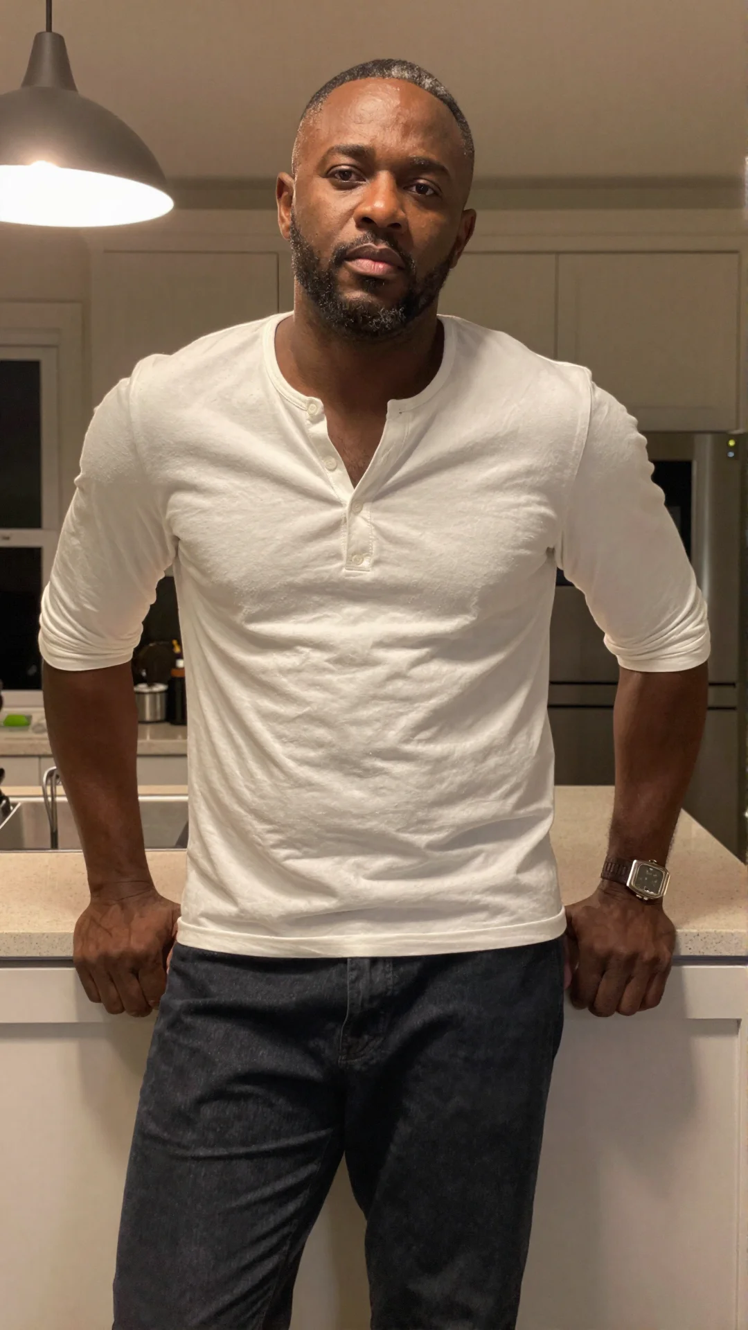 Serious Nigerian man in an unbuttoned white henley leaning on a kitchen counter at night