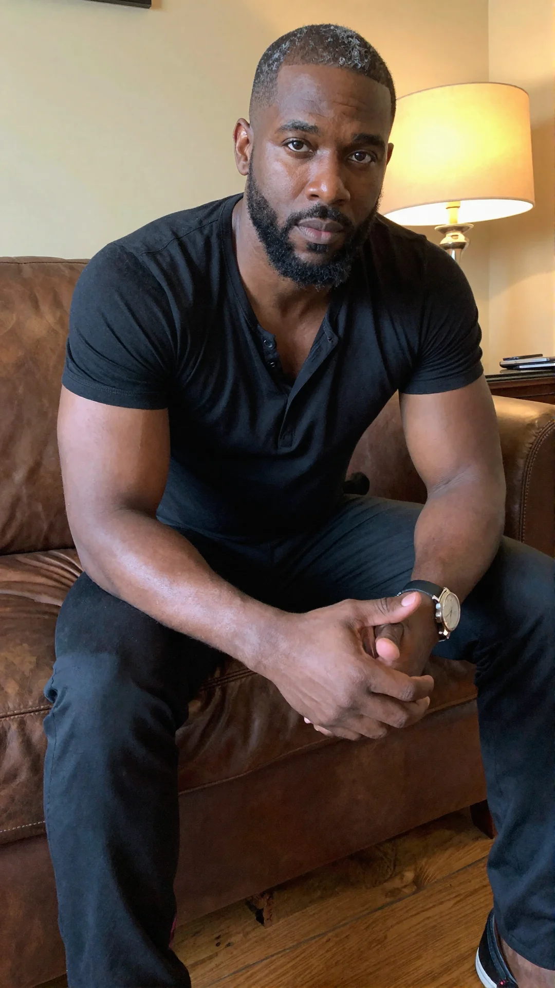 Serious Black American man in an unbuttoned black henley leaning forward on a leather couch